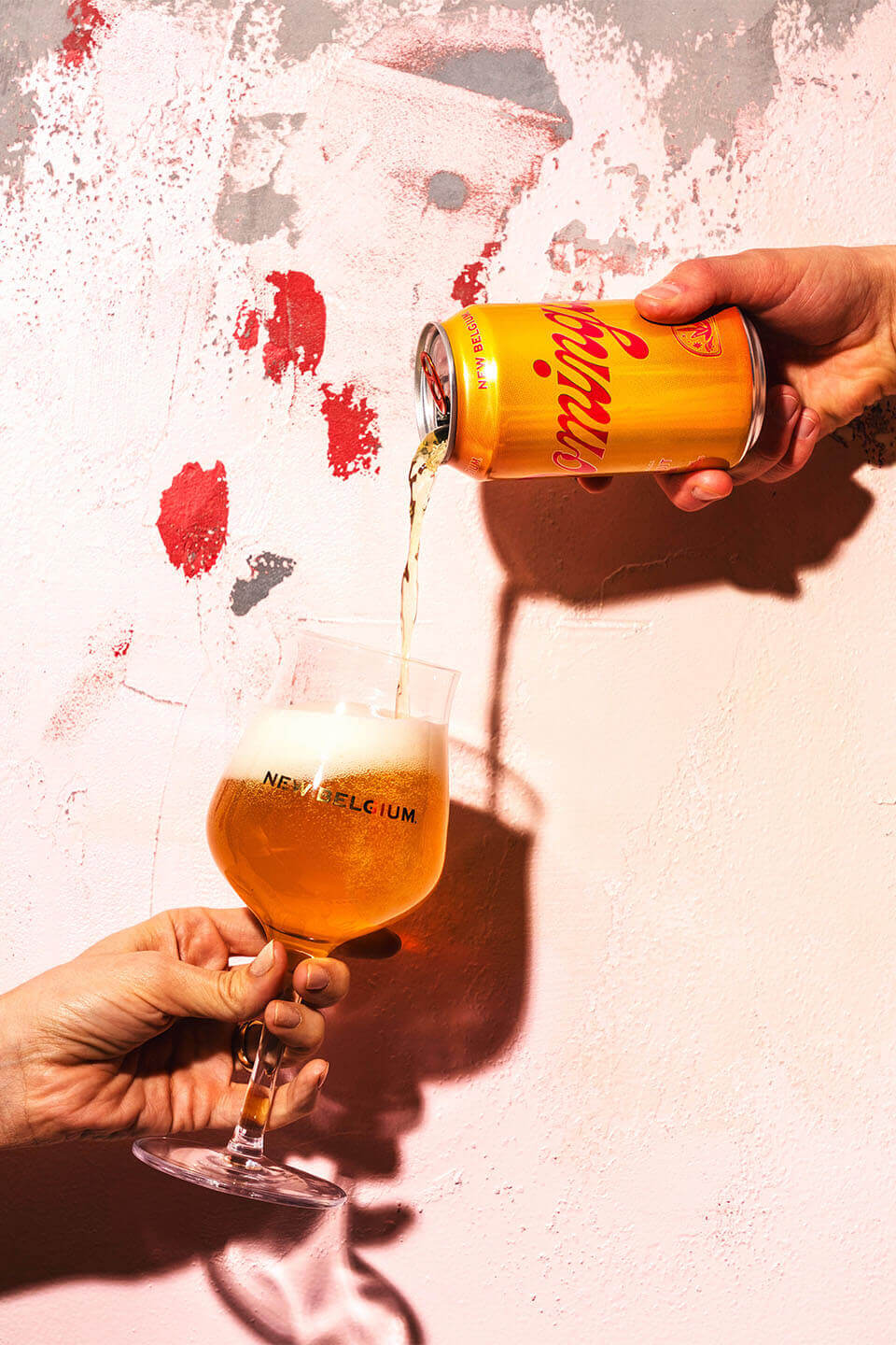 Hand holding New Belgium specialty beer glass while other hand pours can of Dominga into the glass, against white wall with sporadic spots of red. 