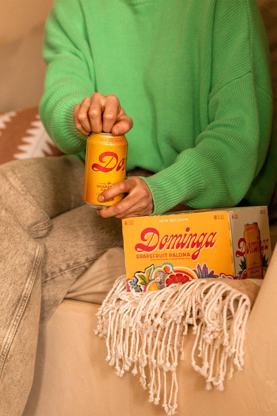 Person sitting on a couch, cracking open a can of Dominga with a 6-pack next to them on the couch. 