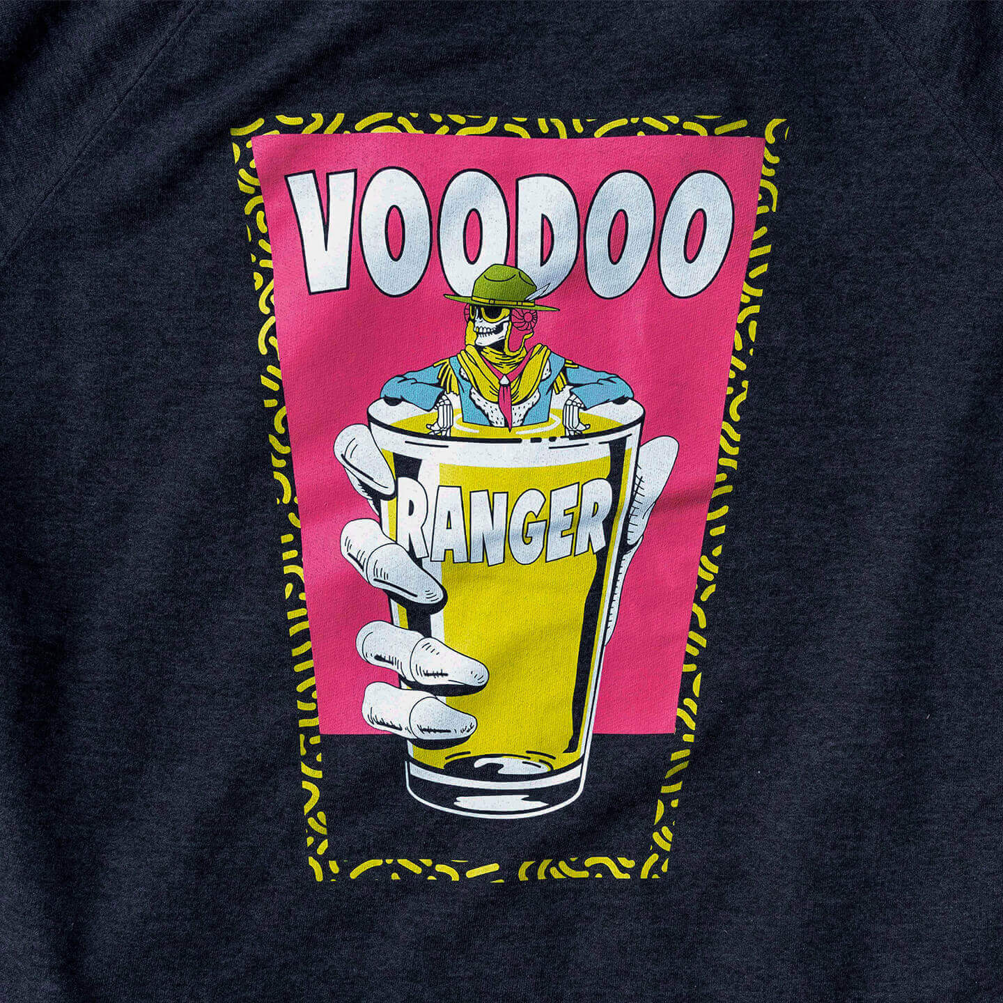 Close-up of laser printed, illustration on the back. It's a pint glass shape with a yellow patterned outline, filled with hot pink three quarters of the way down, and grey the remaining quarter. A large Voodoo hand holds a smaller pint glass filled with yellow liquid, and Voodoo Ranger is soaking in the glass with his arms resting on the side of the pint glass. The word "Voodoo" is large behind the Voodoo Ranger, and the word "Ranger" is smaller and printed on the pint glass. 