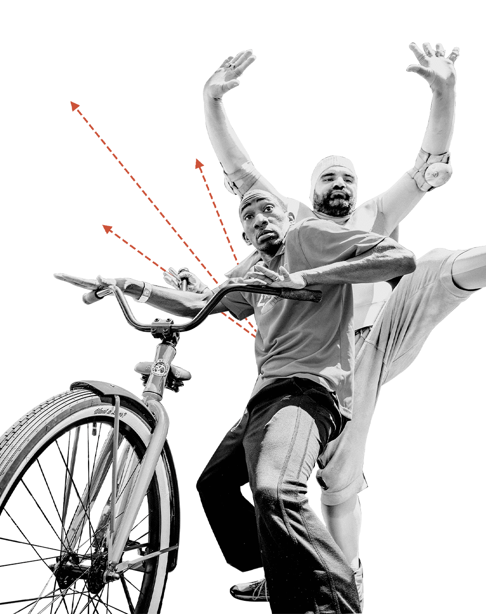 Two people goofing around, one holding onto a bicycle and the other kicking a leg up with his hands in the air. 